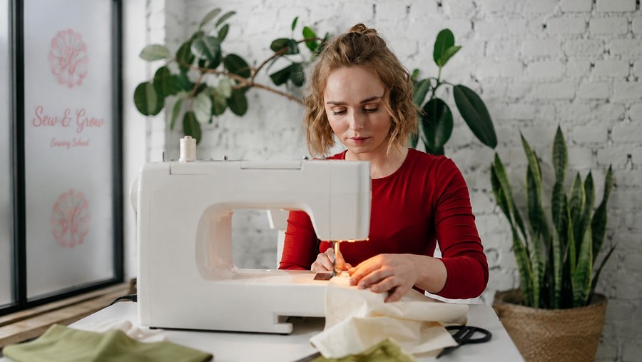 A woman sewing at her machine in a bright, plant-filled studio
